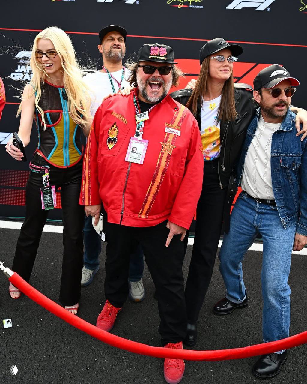 Несколько актёров в яркой одежде и кепках на фоне бренд‑волла Формулы‑1, некоторые улыбаются и машут публике у ограждения