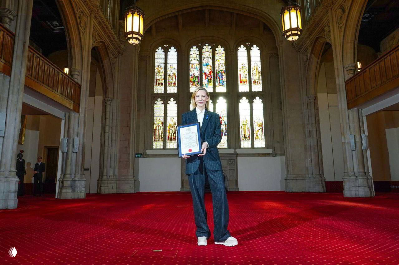 Кейт Бланшетт стоит в большом зале с красным ковром и высокими витражными окнами, держит награду Freedom of the City; общий план.