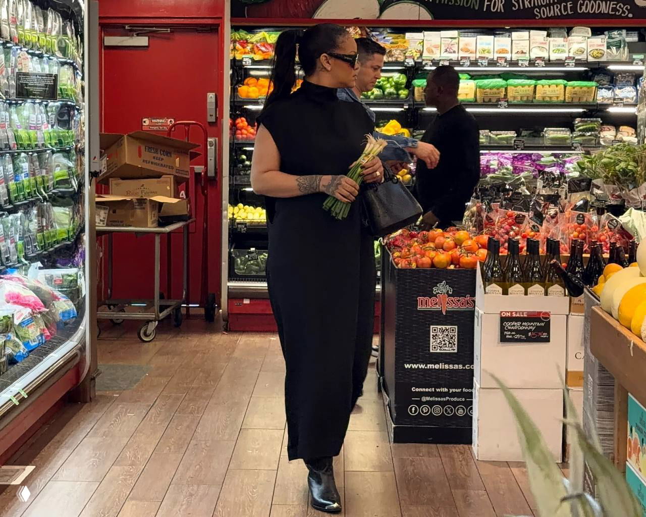 Женщина в черном платье и ботинках идёт по деревянному полу супермаркета, в руках у неё букет зелёного лука и сумка, на фоне витрины с фруктами.