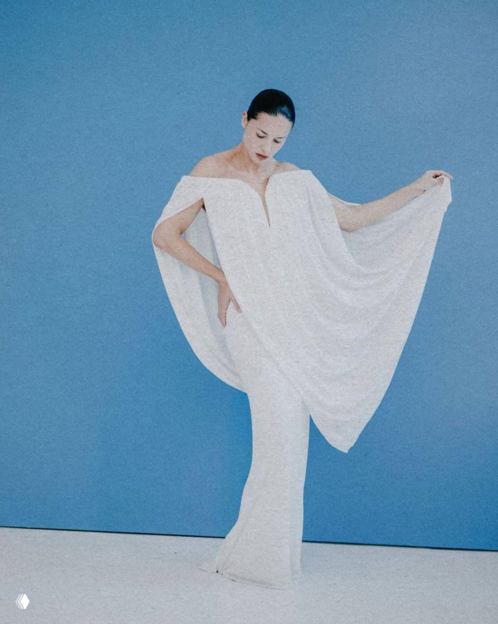 Модель в белоснежном платье, ракурс в полный рост на голубом фоне; воздушная драпировка и образ легкости в кадре.