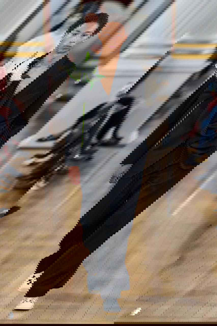 Модель в чёрном драпированном костюме несёт белый цветок — сочетание строгого подбородка и мягкой драпировки на подиуме.