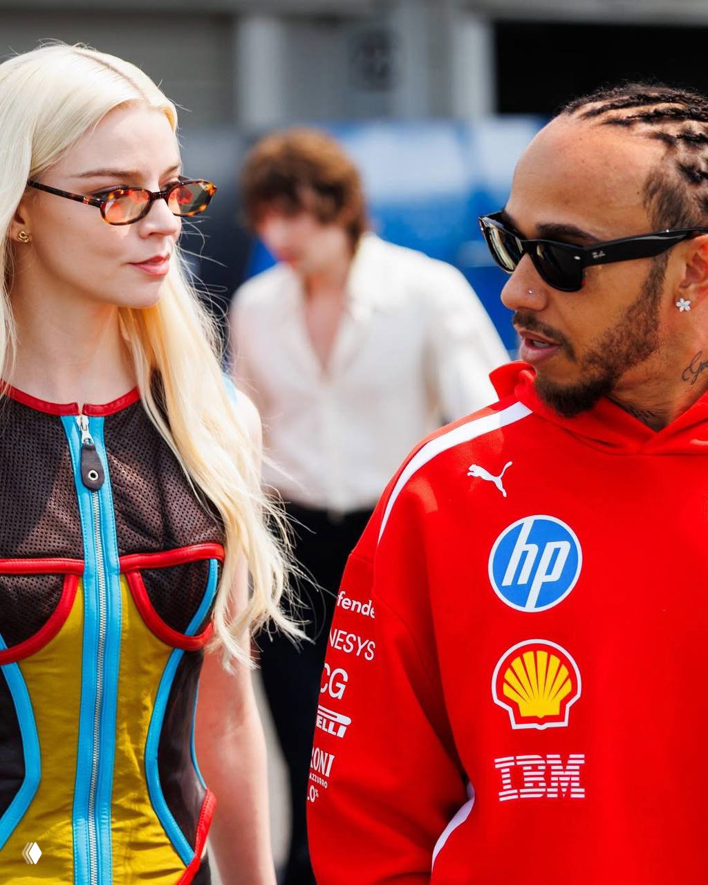 Крупный план двух людей в солнечных очках у гоночной зоны — один в яркой спортивной толстовке с логотипами, другой в лёгком образе, разговор в окружении людей