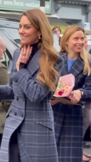 Крупный план: принцесса Кейт в клетчатом пальто улыбается, рядом зрители дарят цветы и фотографируются на фоне городской улицы.