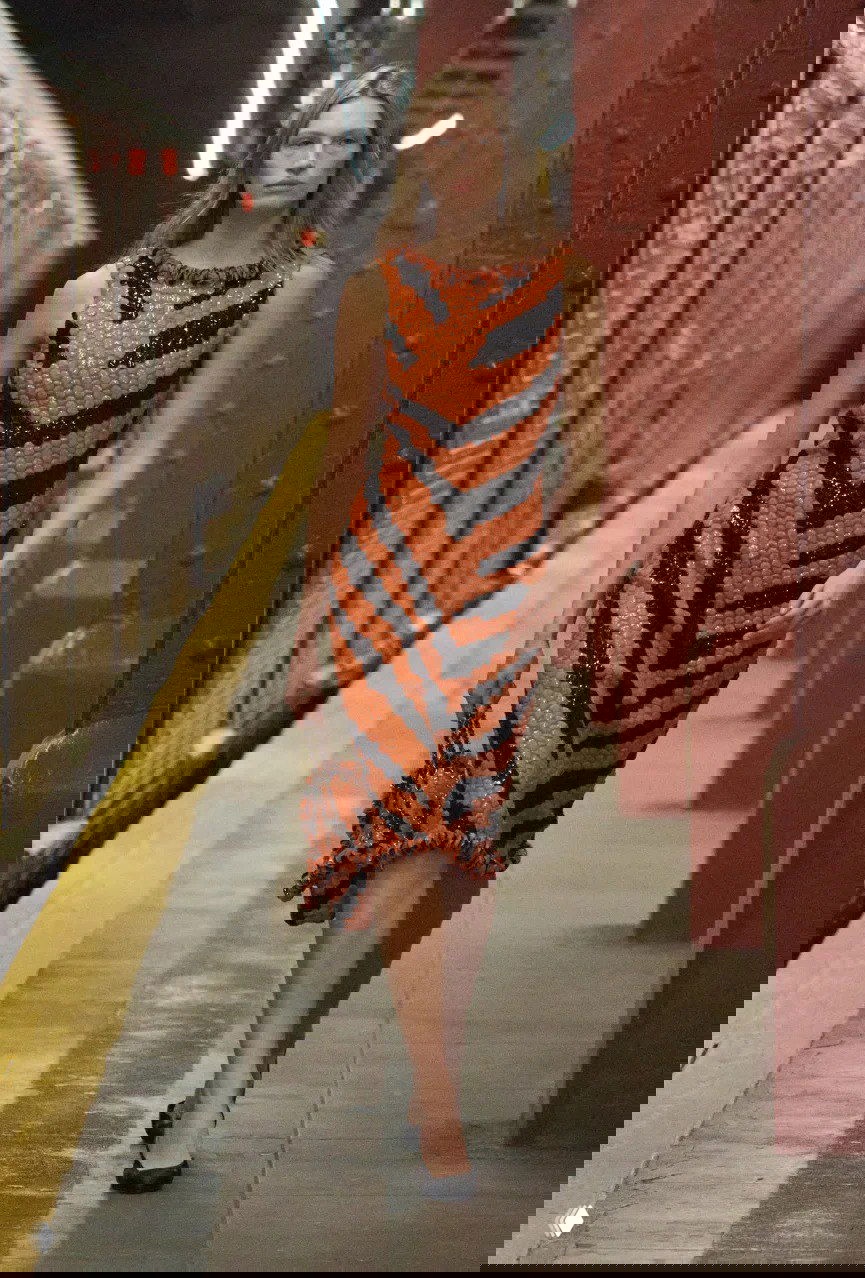 Модель в текстурированном оранжево‑чёрном платье с зебровым узором идёт по платформе метро на показе Chanel, подчёркнуты силуэт и фактура.