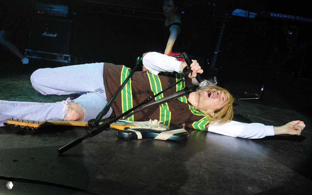 Исполнитель лежит на сцене рядом с электрогитарой в рваных джинсах и полосатой футболке — динамичный и неформальный концертный момент.