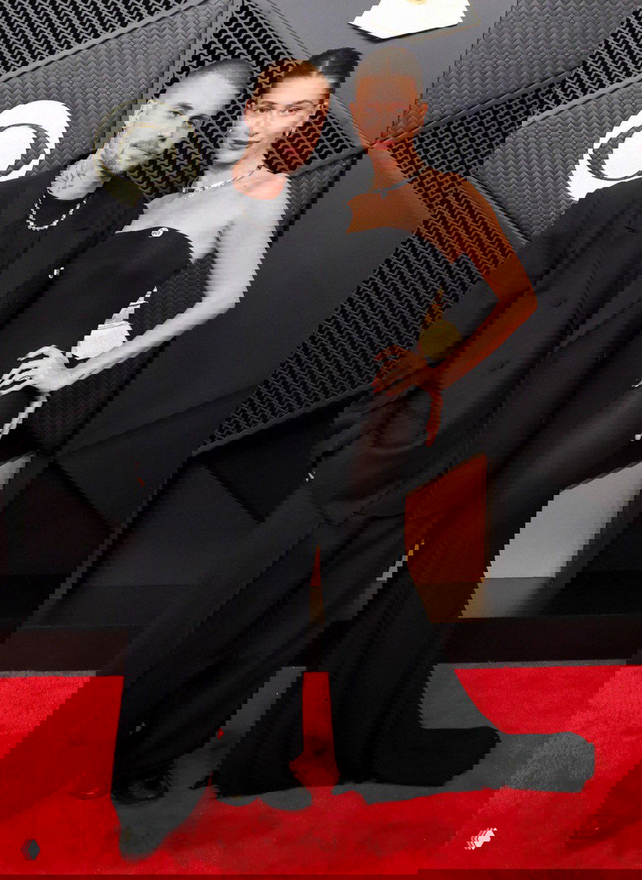 Два человека в элегантных чёрных образах позируют на красной дорожке, минималистичный дуэтный лук на фоне декора и наград церемонии.