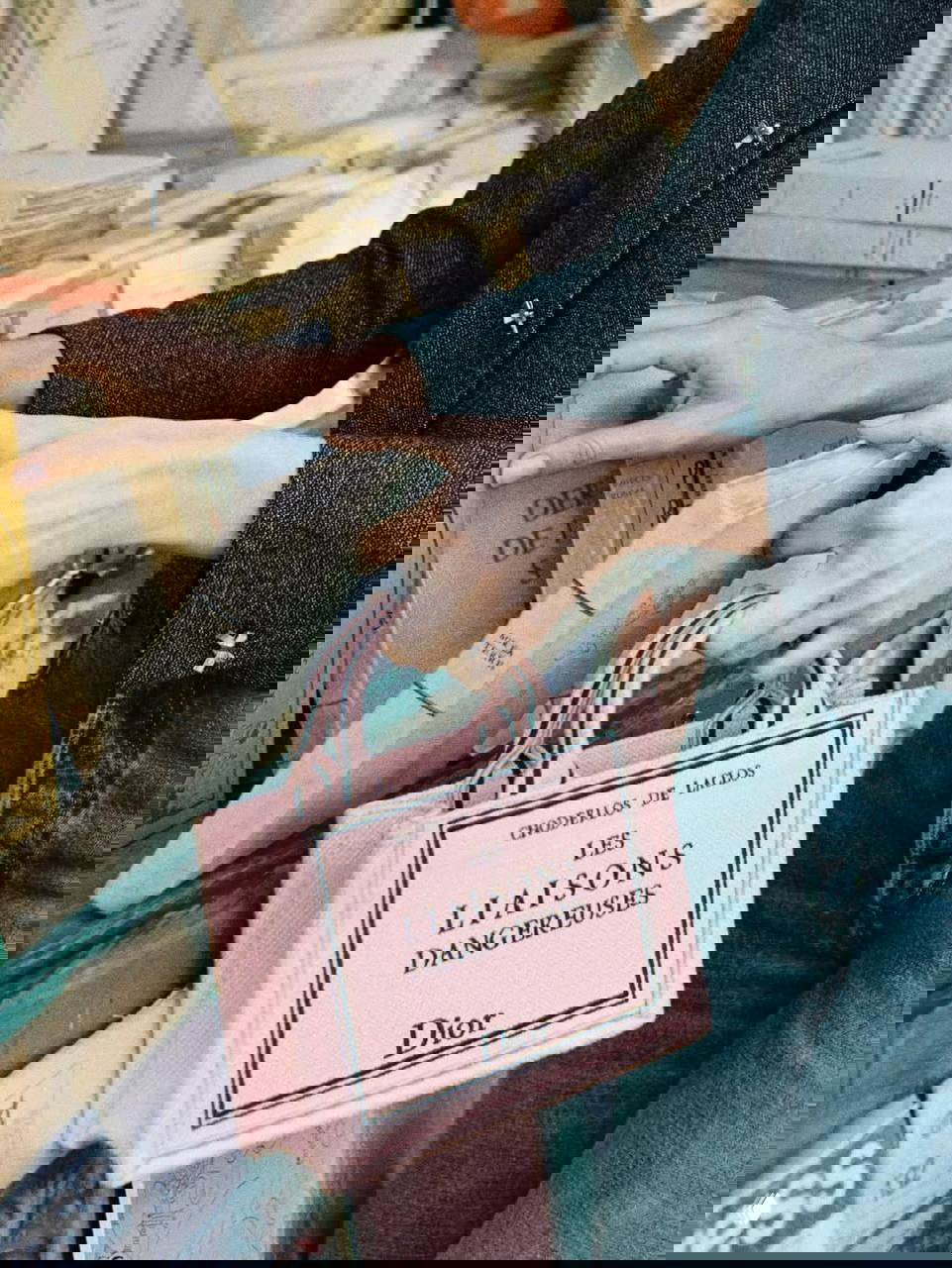 Крупный план розовой сумки Dior с принтом обложки книги «Les Liaisons Dangereuses» на фоне стопок старых томов в книжной лавке под открытым небом.