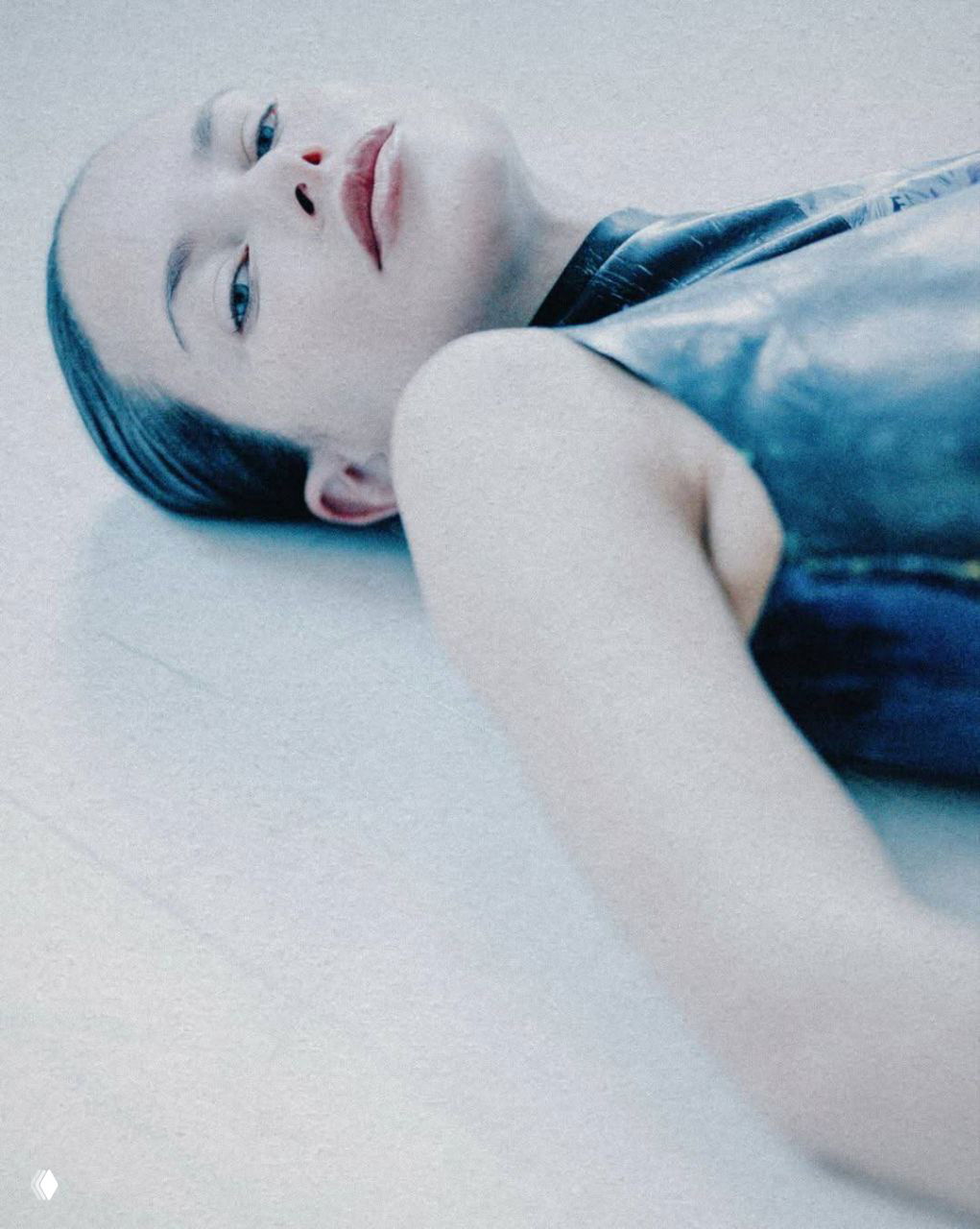 Крупный план модели, лёжа на светлой поверхности, мягкая пастельная палитра и акцент на чертах лица и текстуре кожи.