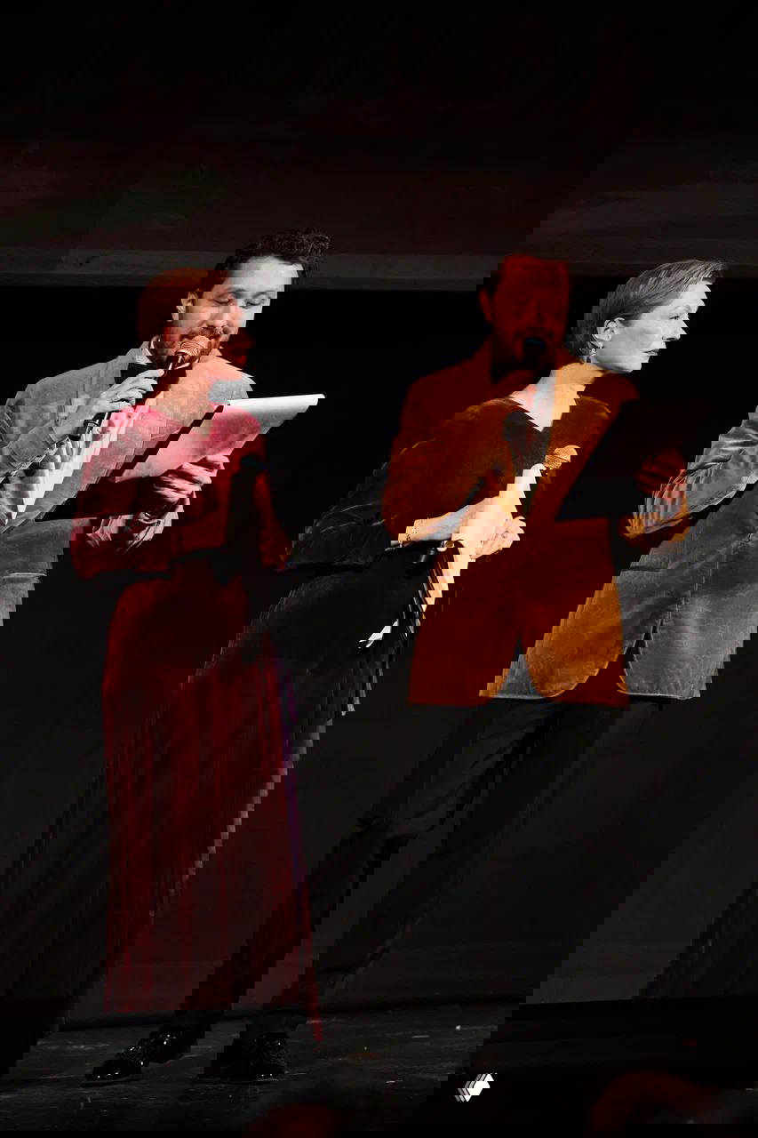 Ведущие церемонии на сцене: женщина в вечернем платье и мужчина с микрофоном и листом с текстом — кадр с момента награждения.