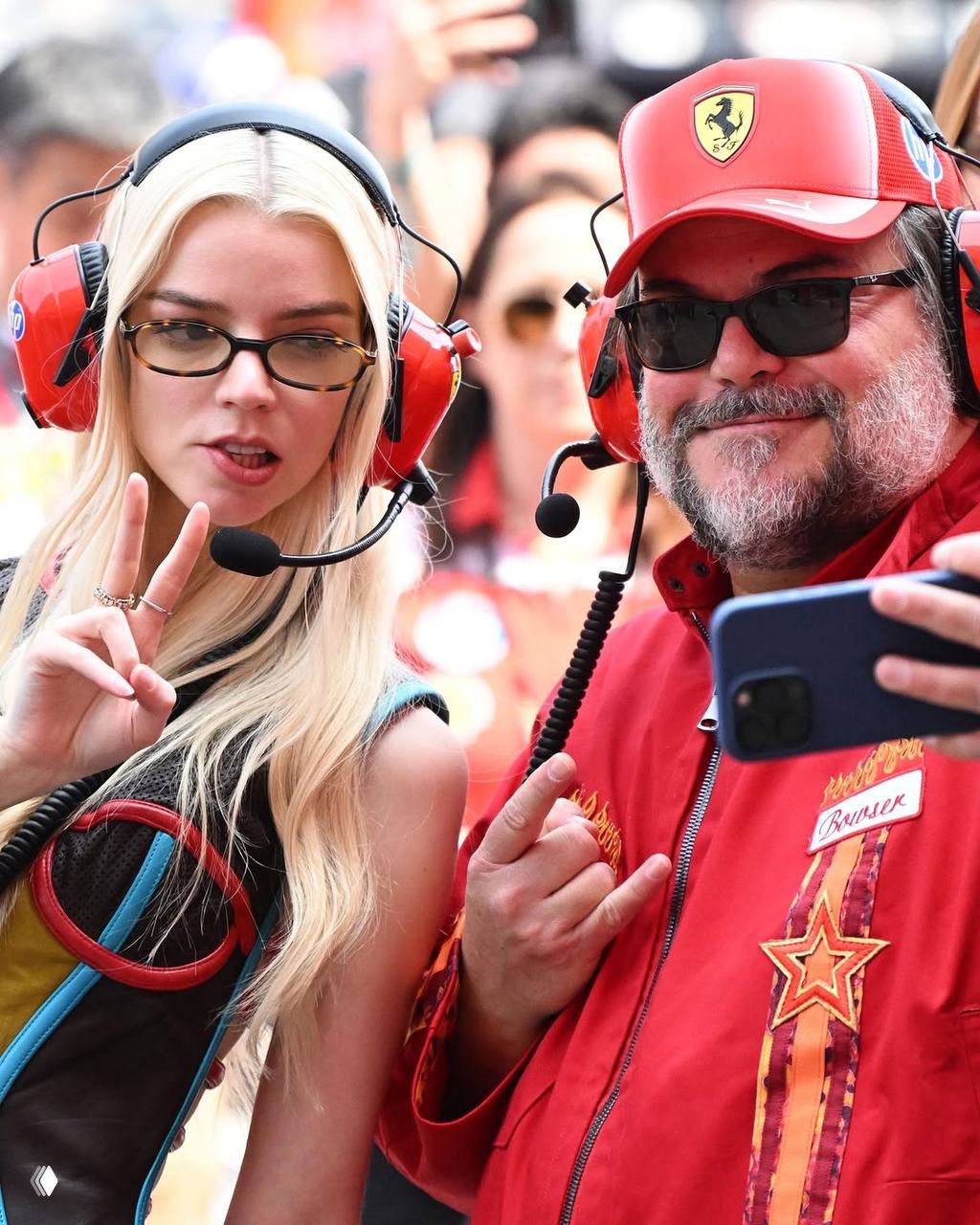 Дама в наушниках для моторных видов спорта и мужчина в красной куртке с микрофоном позируют для селфи и жестов на фоне гоночного стенда