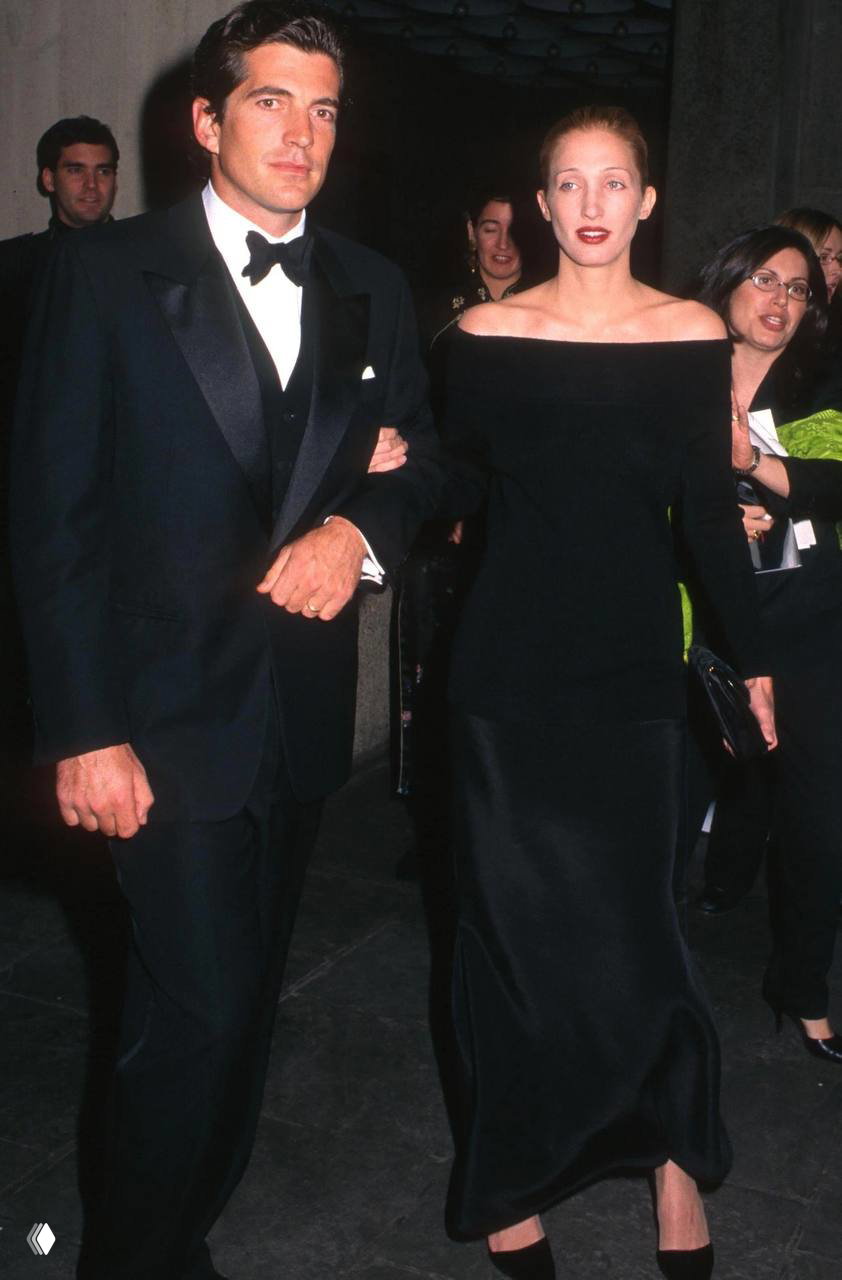 Элегантная женщина в длинном черном платье с открытыми плечами проходит по красной дорожке рядом с мужчиной в смокинге, официальный фон