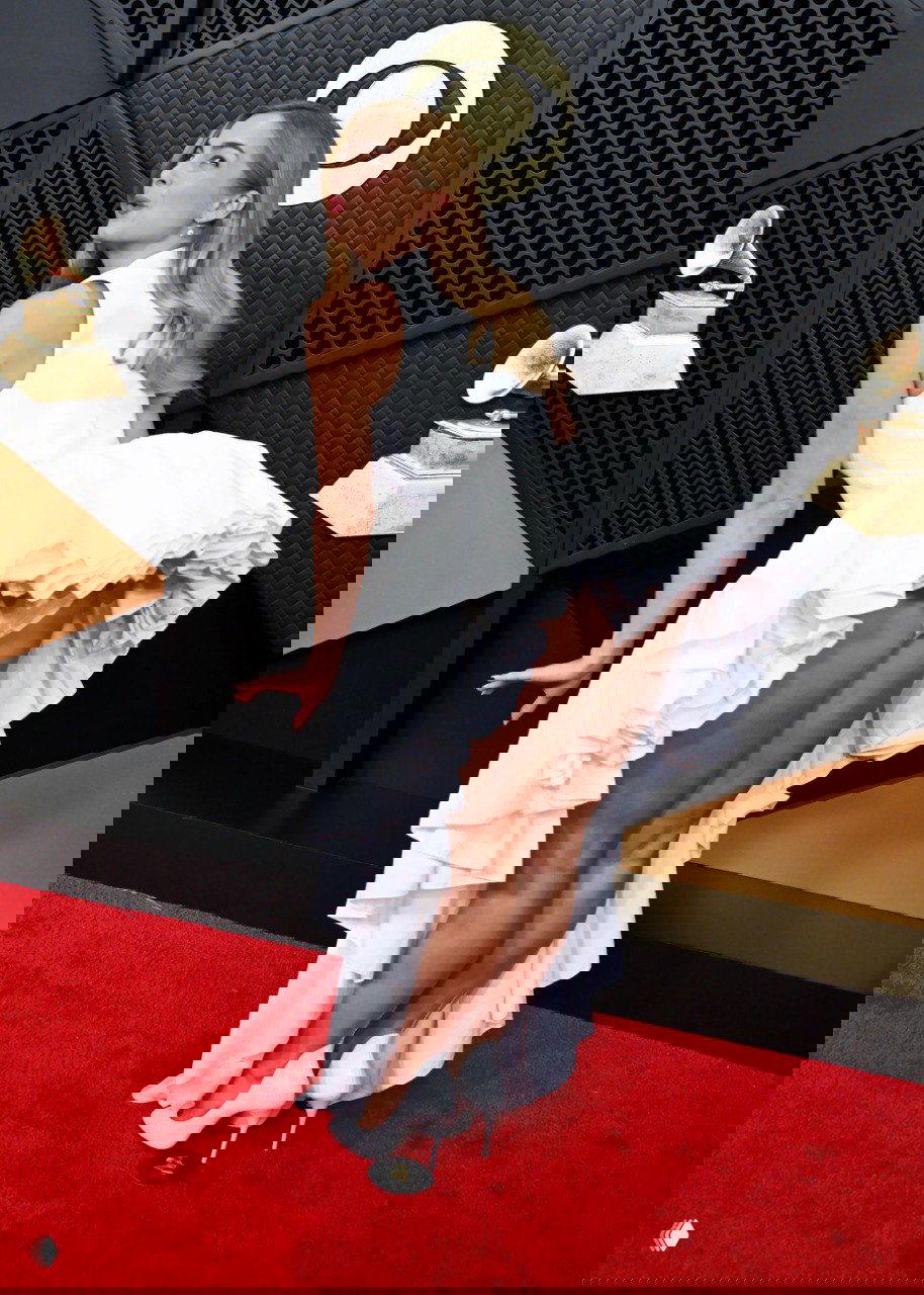 Динамичная поза в белом платье с пышной юбкой на красной дорожке, эффектный момент с приподнятой частью наряда и классические туфли на каблуке.