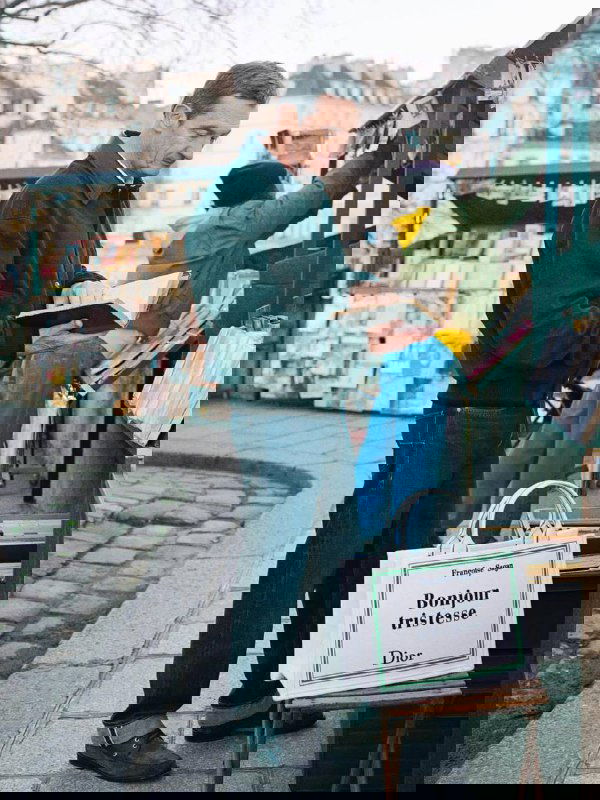 Мужчина просматривает книги у прилавка, рядом стоит белая сумка Dior с надписью «Bonjour tristesse», сцена уличного книжного рынка и аксессуаров бренда.