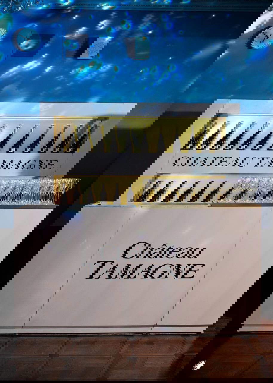 Барная стойка с надписью CHATEAU TAMAGNE и рядом ряды бокалов, оформленная зона напитков для гостей вечернего приёма.