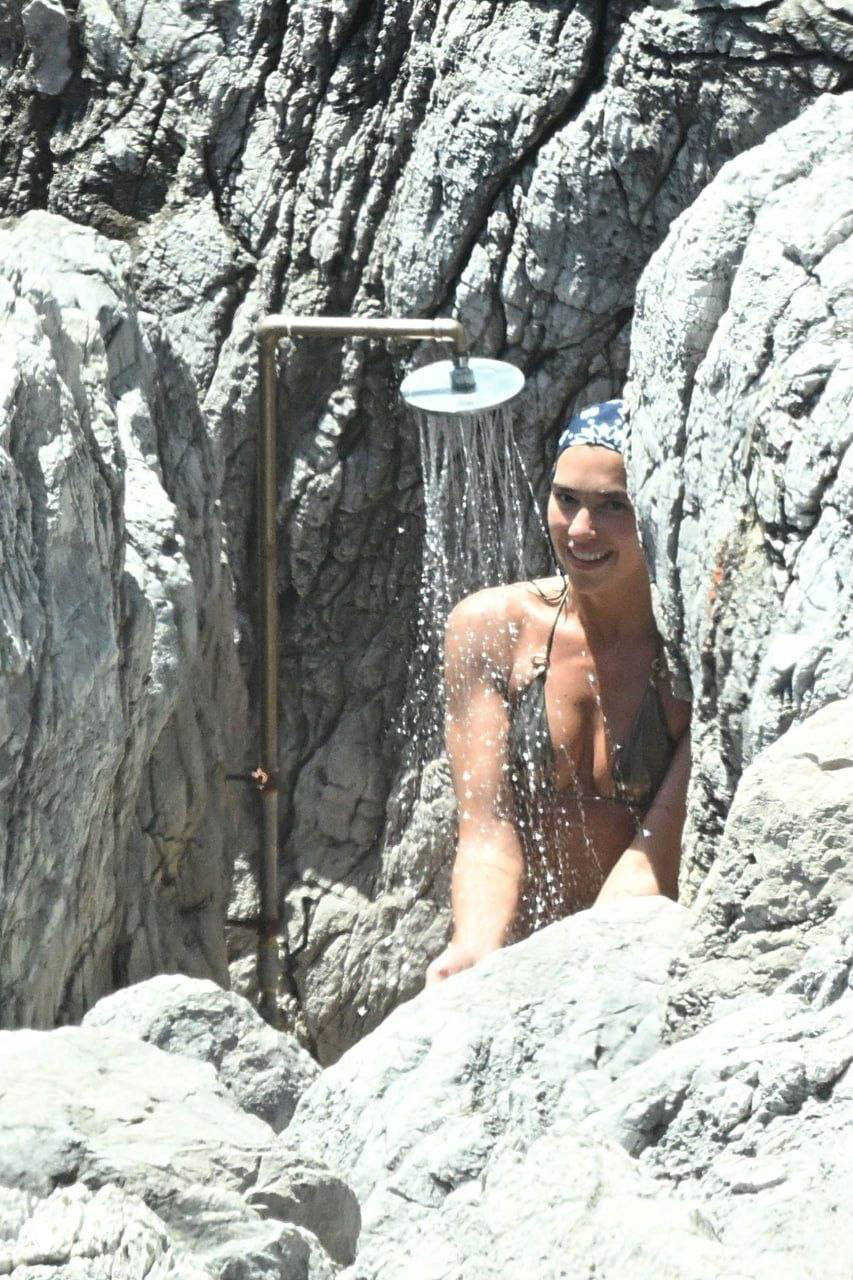Женщина под уличным душем между скалами: купальный костюм, струя воды и каменистая береговая линия, типичный вид укромного места на Амальфи, Салерно.