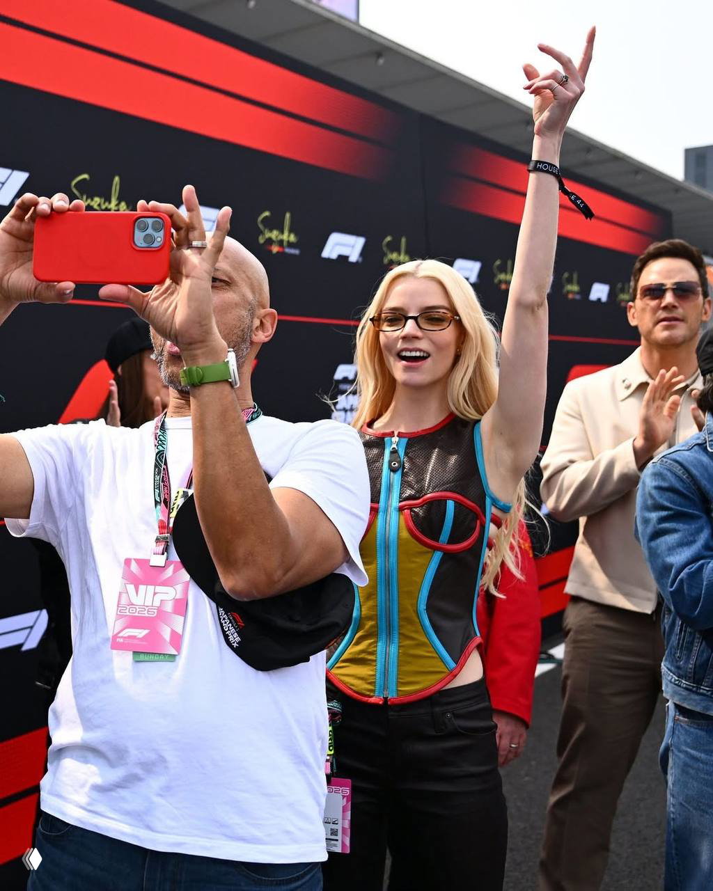 Достойно снятый кадр: женщина с поднятой рукой и мужчина с телефоном делают фото на фоне красно‑чёрного баннера Формулы‑1 в Сузуке