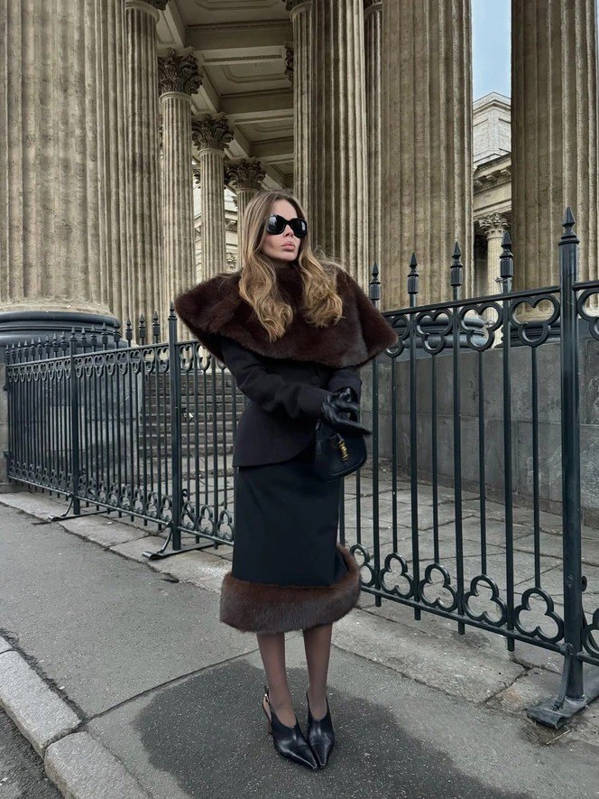 Модель в элегантном тёмном наряде с крупным меховым кейпом позирует у колонн театрального фасада, подчёркивая драматичный городской образ.