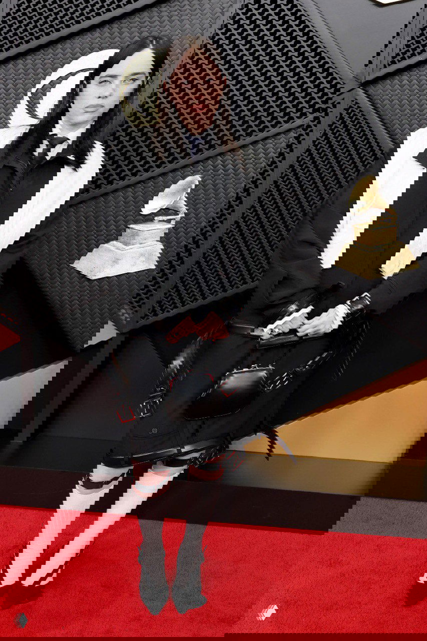 Индивидуальный монохромный образ с жакетом и галстуком на красной дорожке, строгий силуэт, аксессуары в руках и нейтральный макияж.