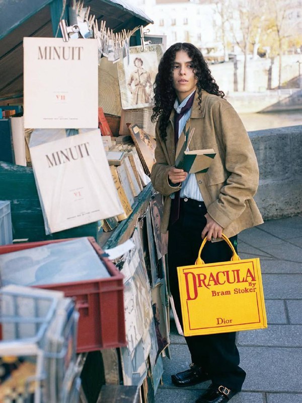 Женщина с жёлтой сумкой Dior «Dracula» стоит у прилавка с книгами; образ напоминает лукбук — сочетание литературы и моды в городской среде.