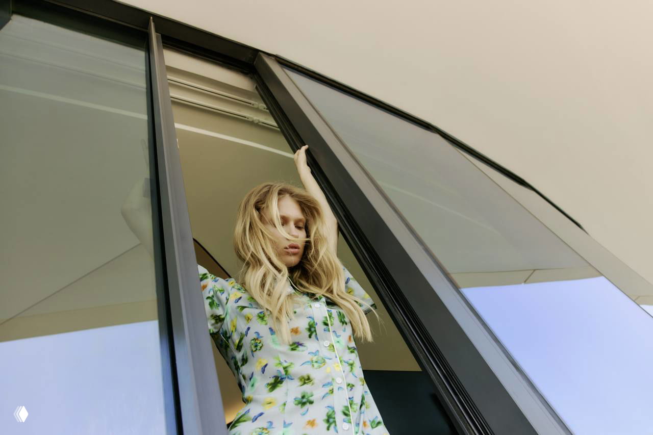 Модель в цветочном платье выходит в окно современного пространства; кадр подчёркивает силуэт, принт и лёгкость летнего образа.