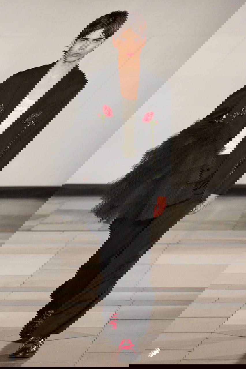 Модель в чёрном пиджаке с тюлевой деталью и красными акцентами на обуви — игра с пропорциями и женственными элементами.