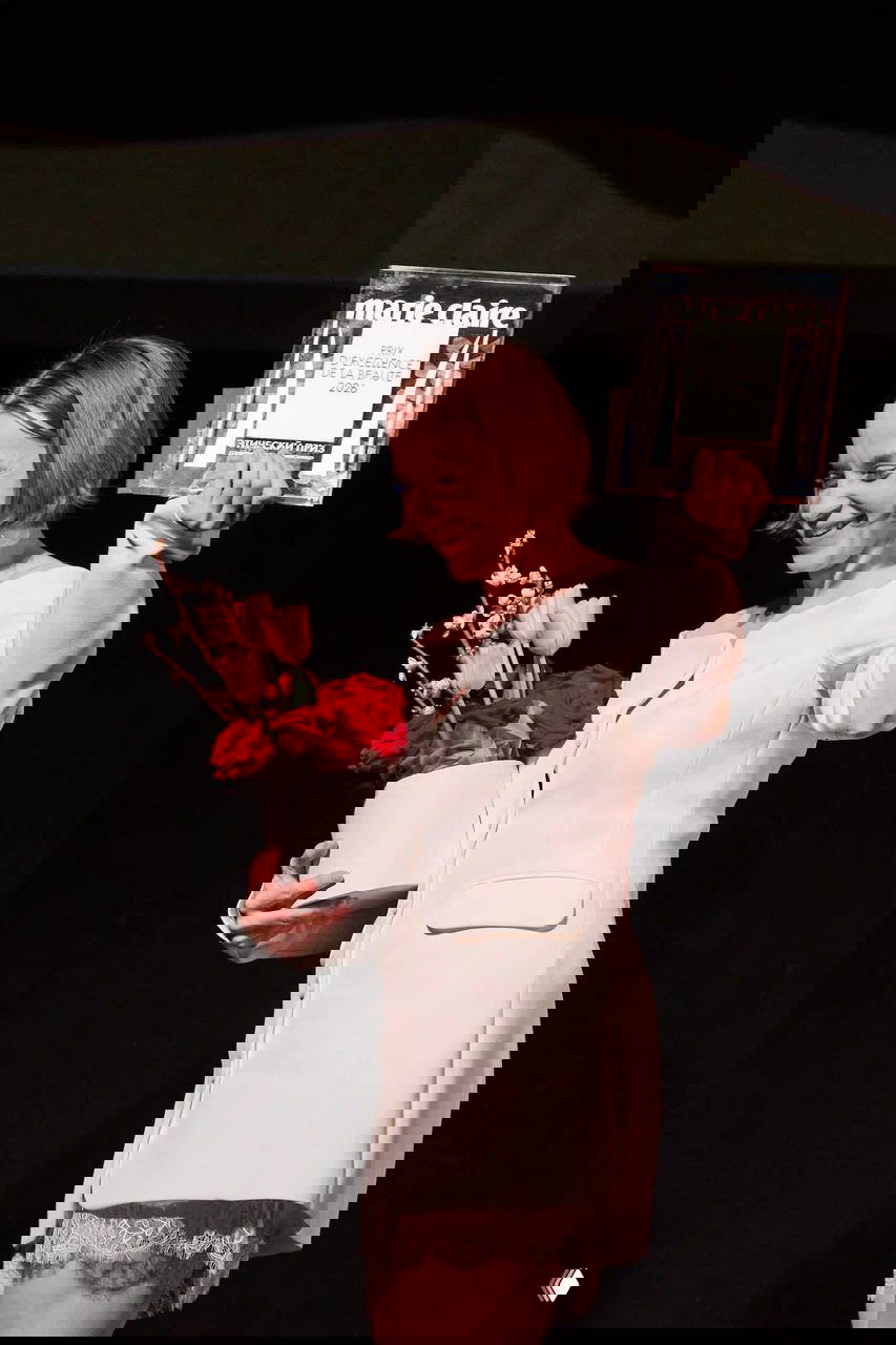 Награждённая держит пластину и букет цветов, улыбается на сцене в светлом платье — момент вручения приза на церемонии.