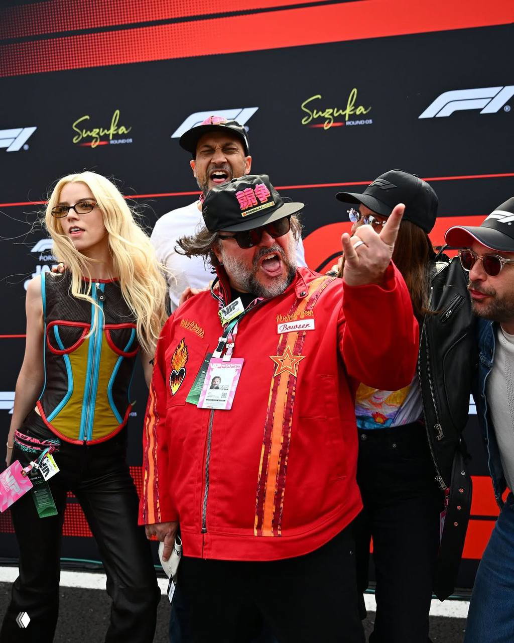 Группа людей на ограждении трассы: один человек изображает движение рукой, рядом — артисты в повседневной одежде и кепках на фоне логотипов Suzuka