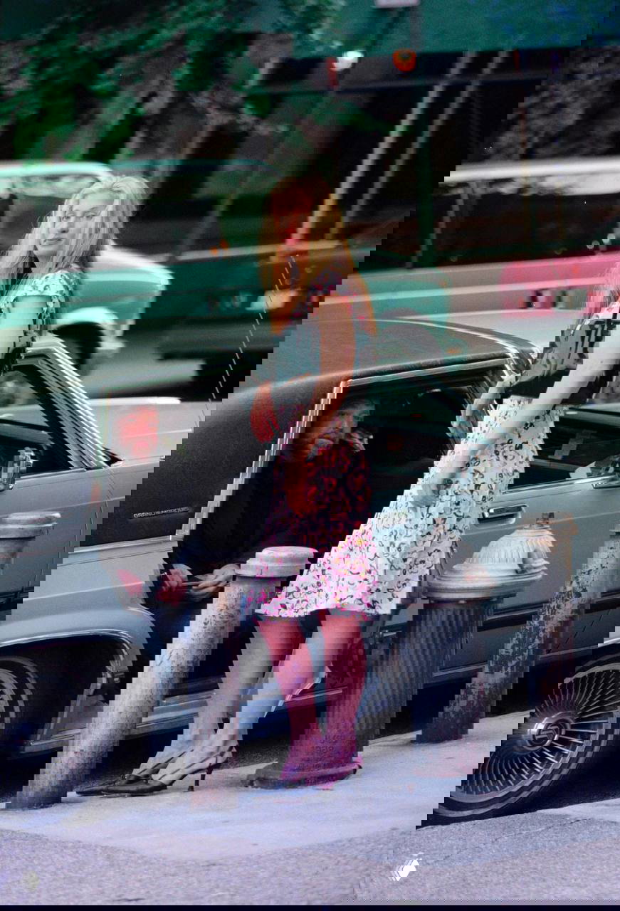 Женщина в летнем платье и туфлях на низком каблуке стоит рядом с автомобилем на городском тротуаре в неформальном кадре у дороги