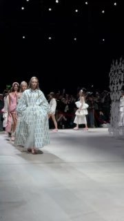 Широкий план подиума: модели проходят между резными белыми панелями‑инсталляциями, на фоне видны кадр и операторы показа в Манеже.