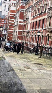 Улица в Лондоне у исторического здания: прохожие и предполагаемая охрана у тротуара на фоне красно-кирпичного фасада и ограждений.