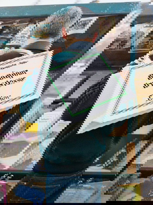 Мужчина читает книгу, у него большая тёмно‑серая сумка Dior с надписью «Les Fleurs du Mal», кадр подчёркивает книжную эстетику коллекции аксессуаров.