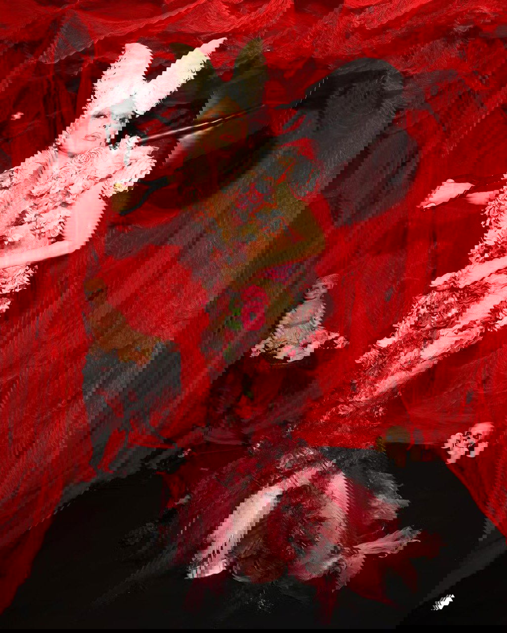 Полный рост Леди Гаги в прозрачном платье с цветочными аппликациями и хвостом из тюля, она держит большую лопату в красном комнате с куклой.