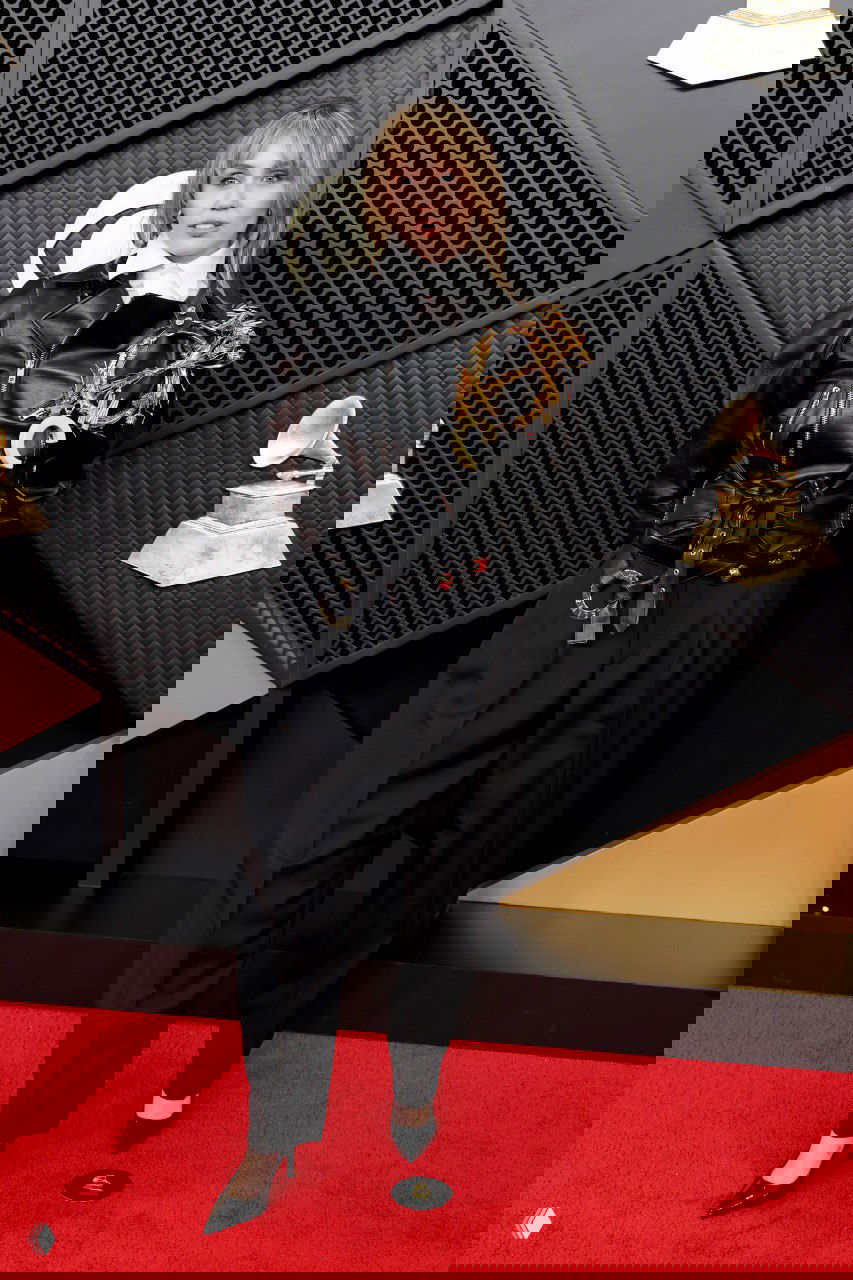 Кэжуал‑рок образ с чёрной кожаной курткой и широкими брюками на красной дорожке, неформальный стиль и выразительный аксессуар на плече.