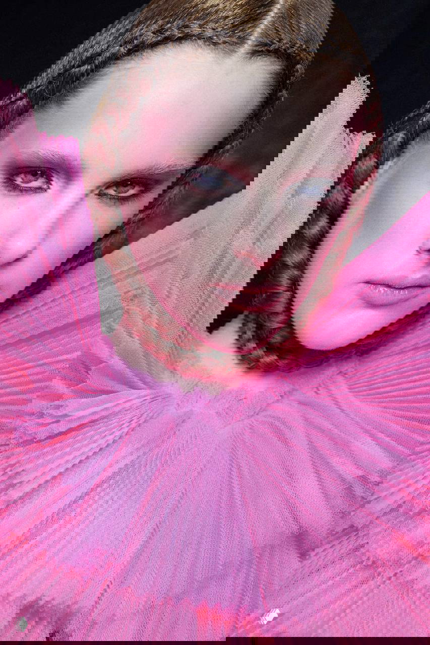 Модель в ярком розовом наряде с выраженным кукольным румянцем и дымчатым акцентом вокруг глаз; фэшн-фотография с сильным цветовым решением.