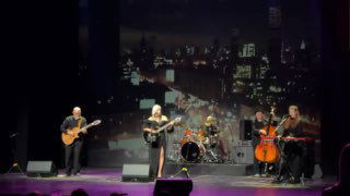 На сцене Алена Свиридова с музыкантами: вокал у микрофона, гитары, виолончель и ударные на фоне ночной городской проекции.