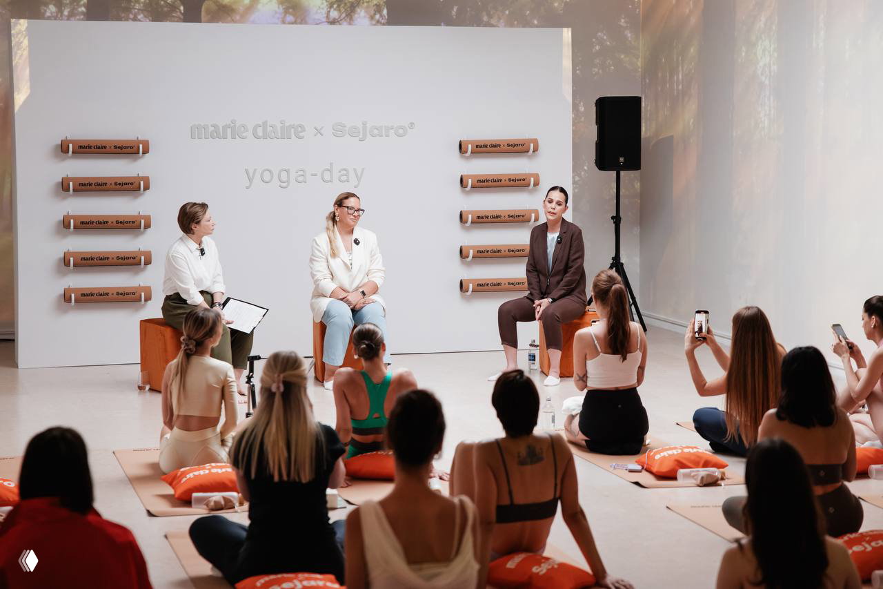 Публичная беседа на сцене Yoga Day: три спикера сидят на креслах перед аудиторией, логотипы Marie Claire и Sejaro на стене.