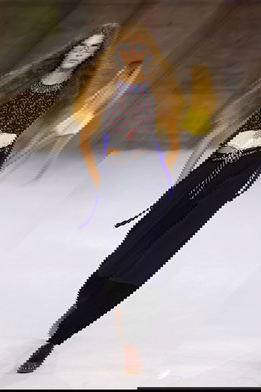 Модель в бохо-образе с коротким цветочным топом, свободными брюками и солнцезащитными очками — свободная эстетика и ретро-волосы на подиуме.