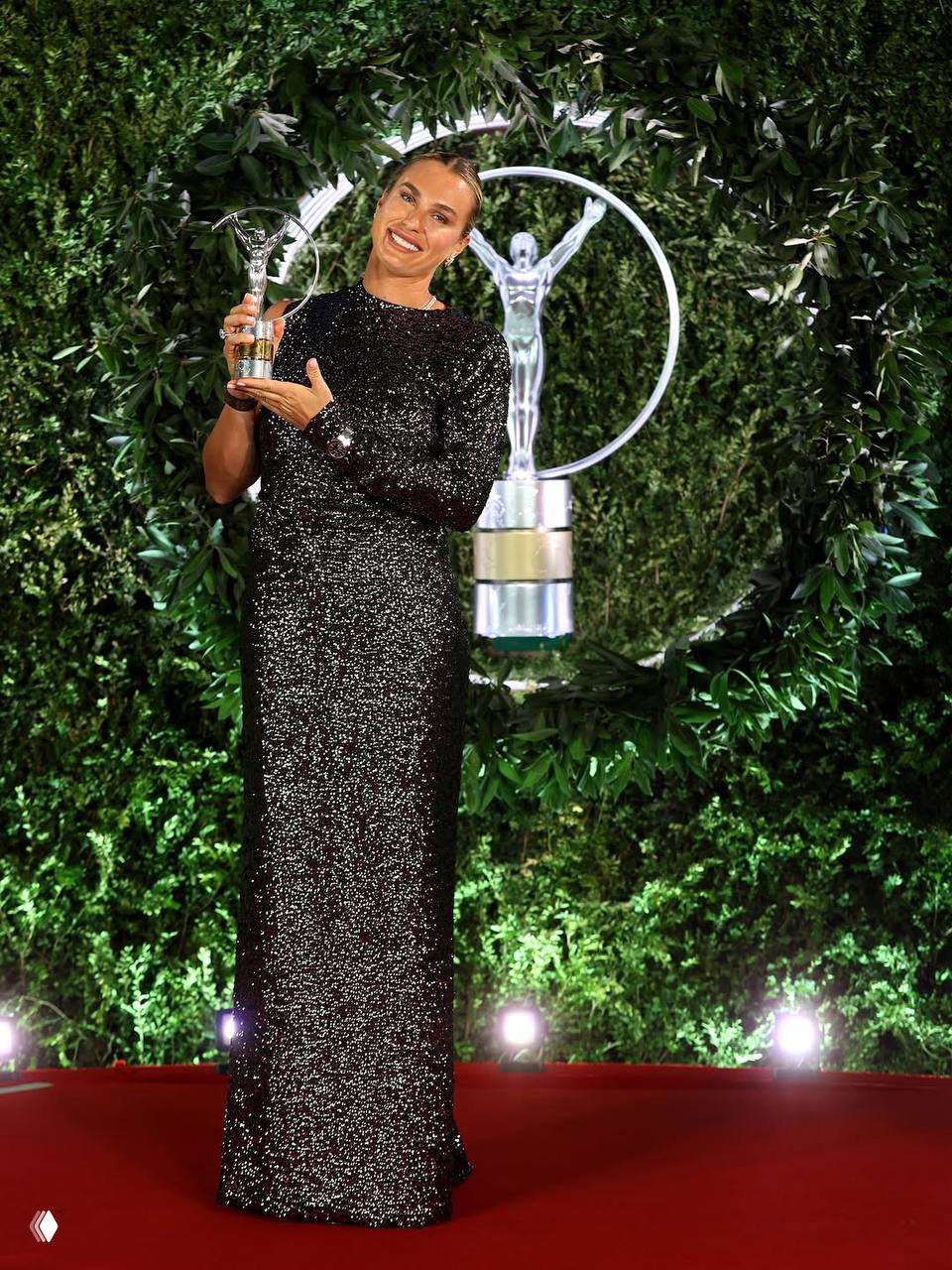 Полноформатный портрет женщины в блестящем платье с той же статуэткой на фоне зелёной декоративной инсталляции на красной дорожке мероприятия.