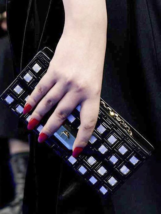 Рука с ярким красным маникюром держит лакированный клатч с геометрическим декором — акцент на форму и текстуру аксессуара.