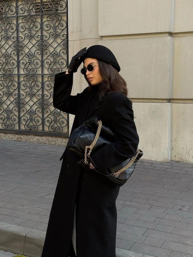 Женщина в тёмной одежде и шляпке-клош с солнцезащитными очками на городской улице — сочетание ретро и современного силуэта.