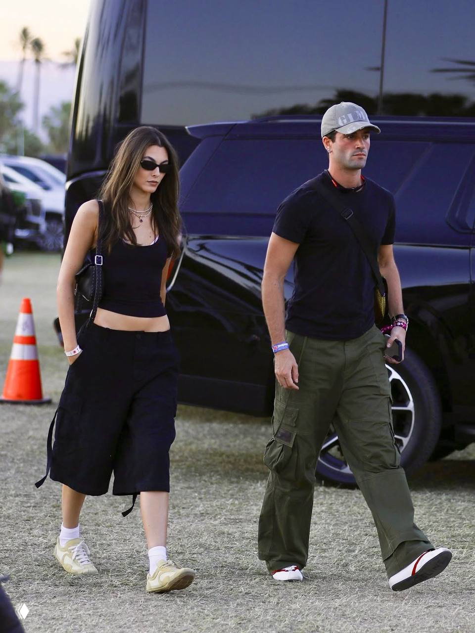 Пара идёт мимо тур‑автобуса: девушка в чёрном топе и укороченных шортах, мужчина в кепке и карго‑брюках — повседневный фестивальный лук.