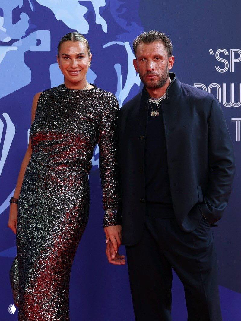 Две фигуры на красной дорожке: женщина в вечернем платье и мужчина в тёмном костюме позируют перед фотозоной мероприятия.