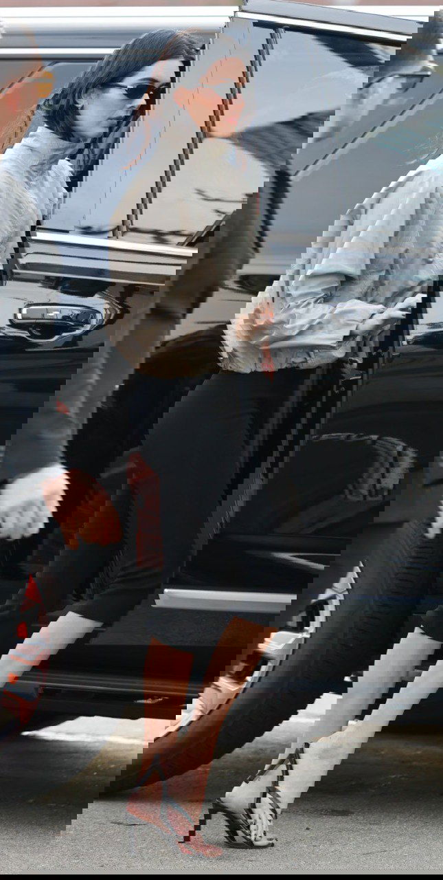 Женщина в бежевой ветровке и чёрных укороченных брюках садится в чёрный внедорожник на парковке у стадиона, кадр в уличном стиле