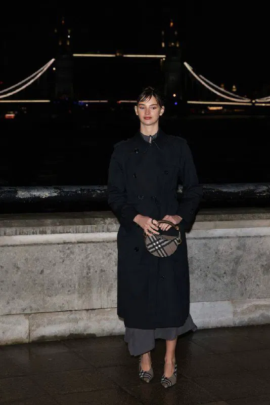 Модель в чёрном тренче и с круглой сумкой с фирменным узором позирует на ночной набережной у моста — городской лаконичный образ.