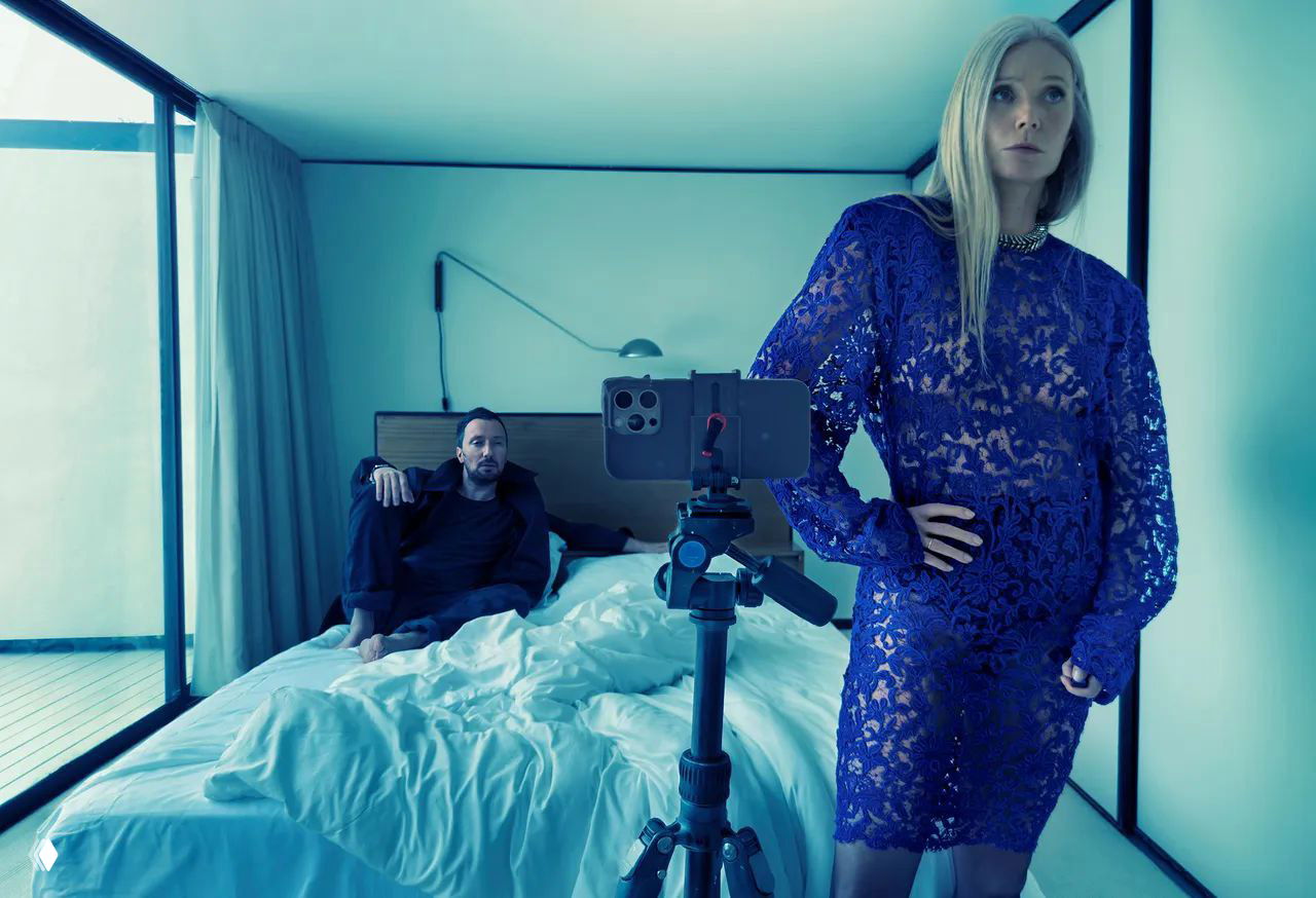 Женщина в синем кружевном платье стоит в спальне перед камерой на штативе; мужчина сидит на кровати, холодная цветокоррекция.