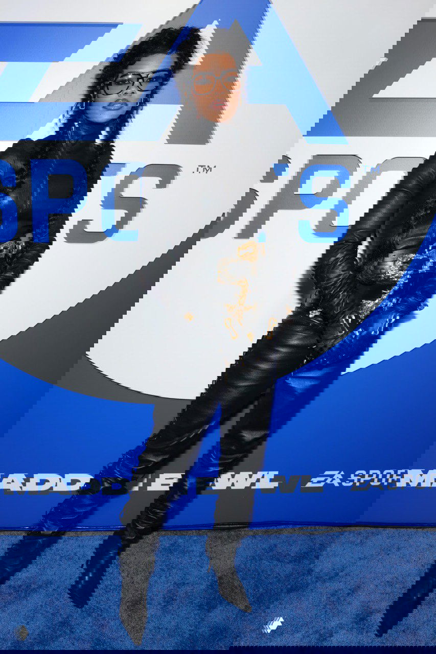 Женщина в чёрном кожаном костюме позирует на синем фоне пресс-волла, образ подчёркнут аксессуарами и модными элементами, виден логотип события