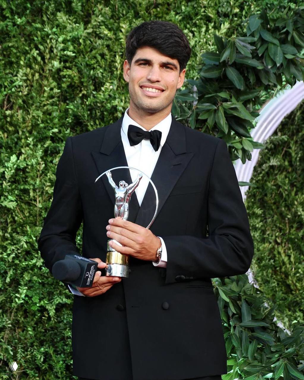 Молодой мужчина в чёрном смокинге держит серебристую статуэтку премии на фоне зелёной декорации и улыбается в камеру.