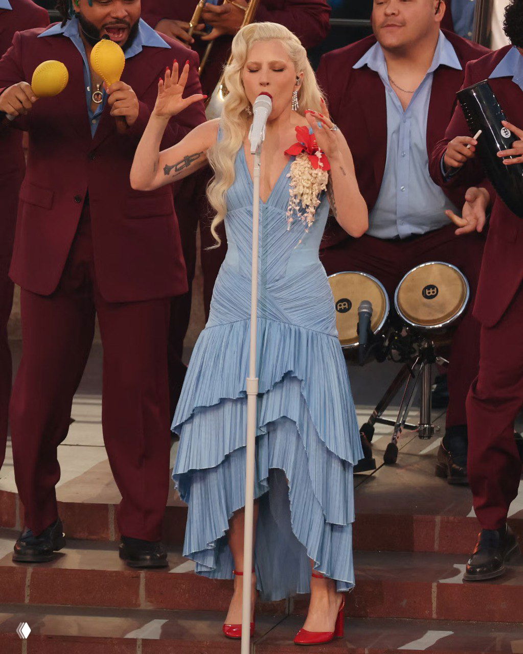 Певица в голубом многоярусном платье поёт на сцене, окружённая музыкантами, кадр сделан во время сценического выступления на стадионе