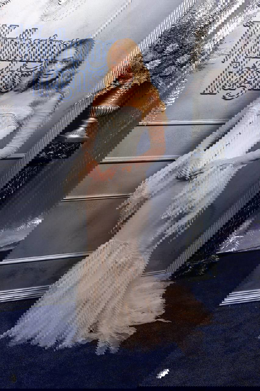 Полный рост актрисы в бежевом корсетном платье с текстурированным низом на фоне логотипа премьеры «Грозового перевала»