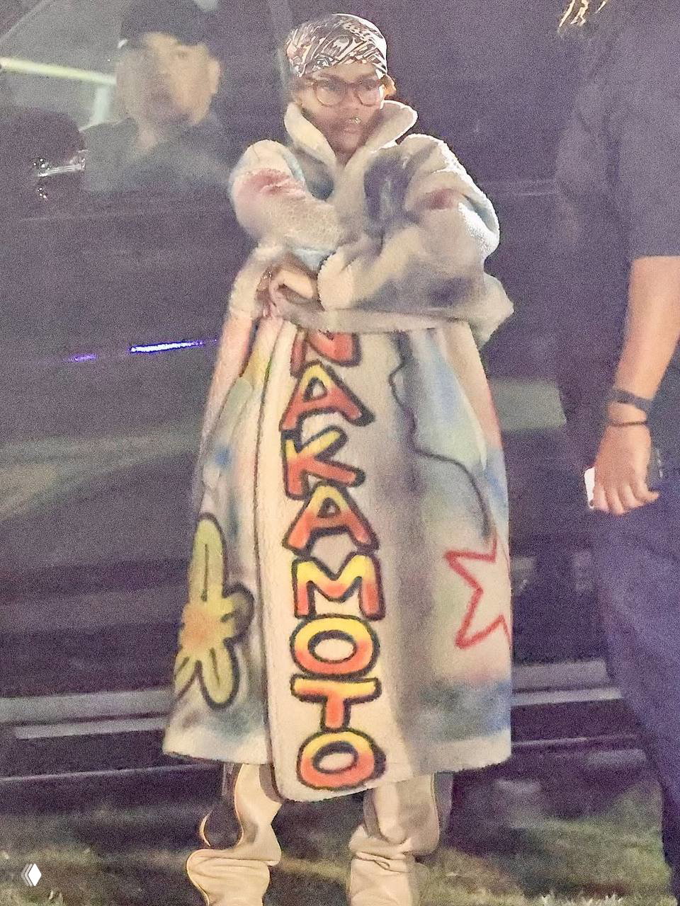 Необычное длинное пальто с граффити‑надписью и ярким принтом на фоне вечернего фестиваля Coachella — экстравагантный сценический образ.